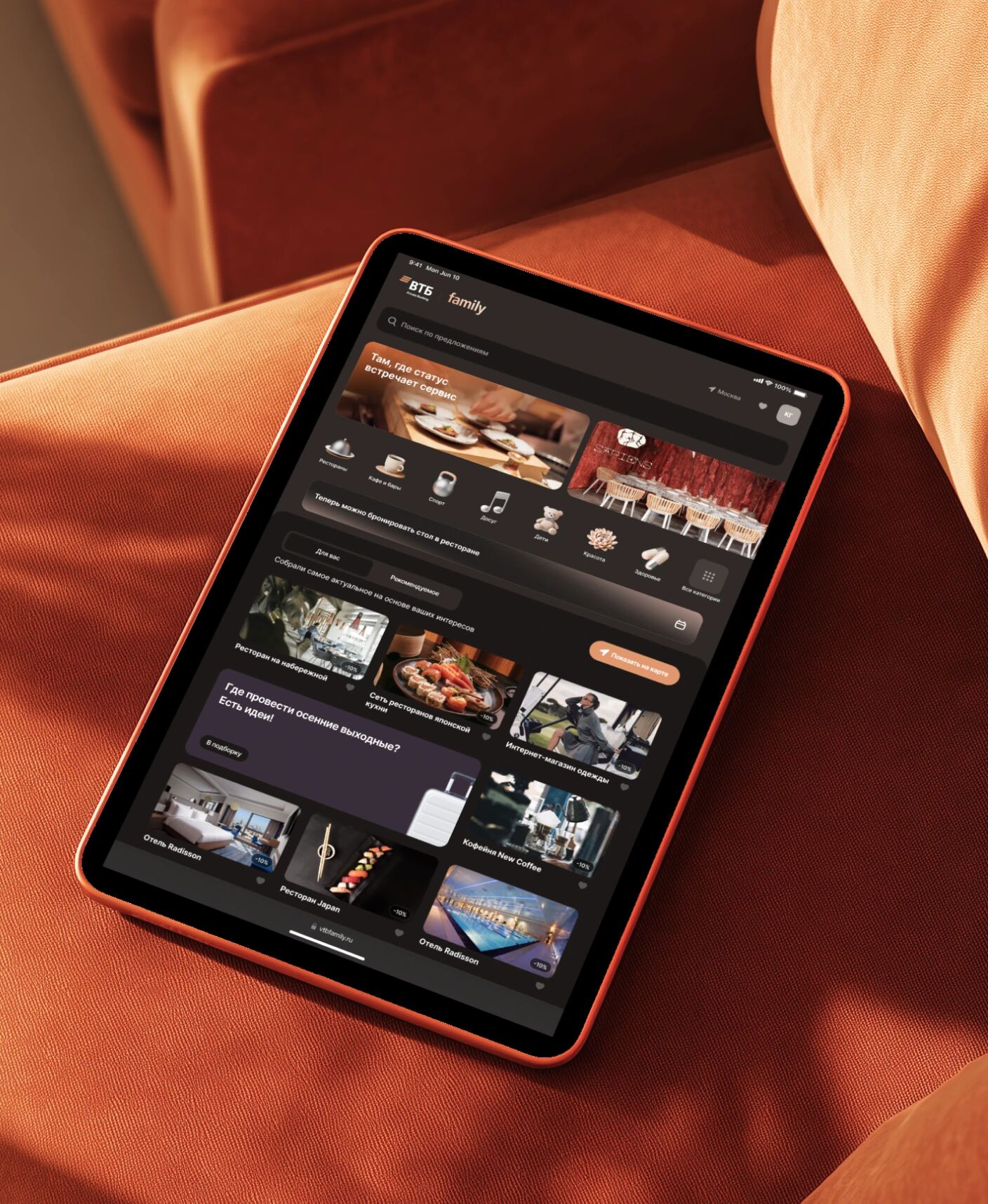 Планшет с тёмным интерфейсом ВТБ Family на оранжевом кресле, главный экран с категориями и персональными рекомендациями